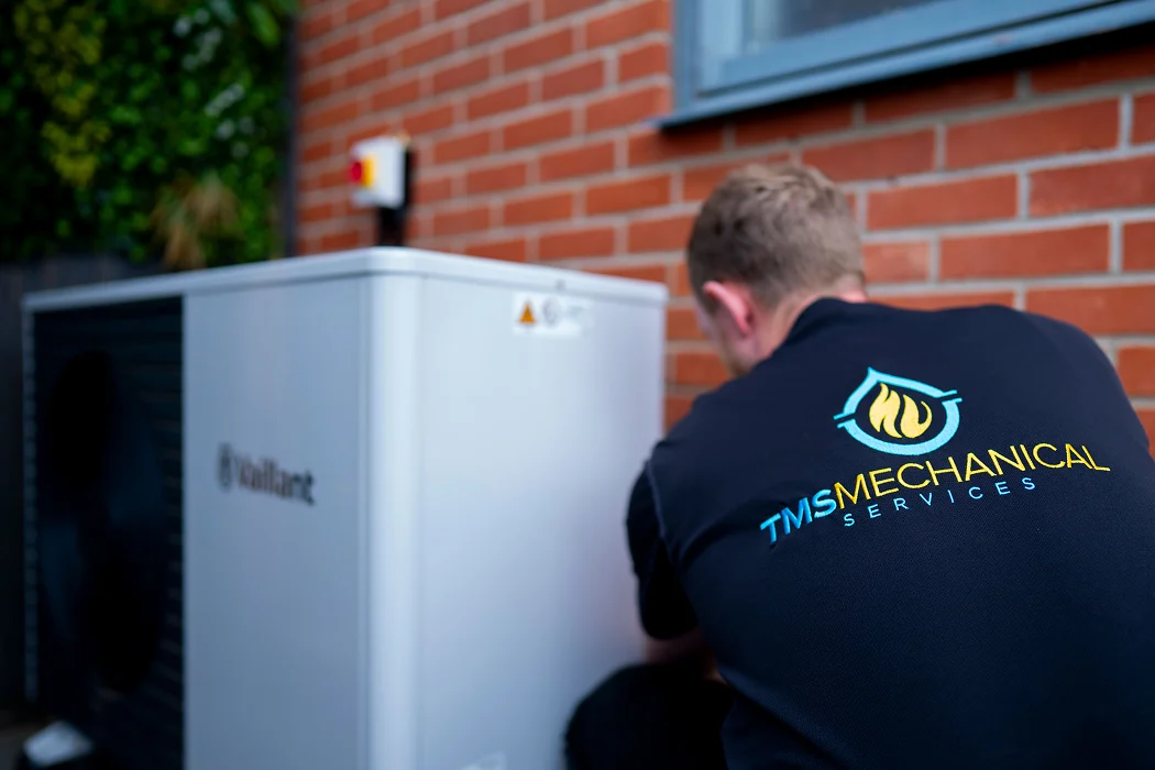 Technician from TMS Mechanical Services installing a Vaillant air source heat pump in a residential setting with brick wall background.
