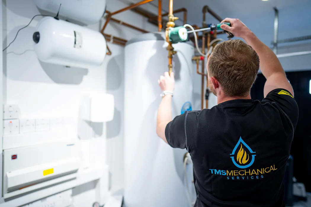 Technician adjusting plumbing equipment in a heating system, featuring TMS Mechanical Services branding, in a modern workspace focused on gas mains commissioning and pressure testing.