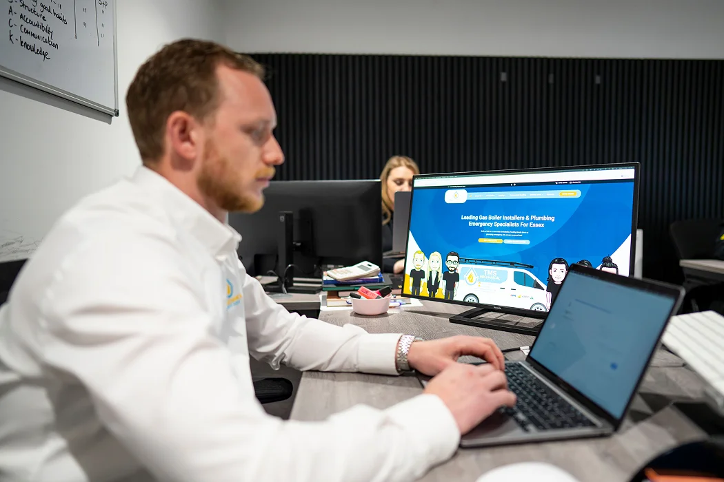 Man working at a desk with a laptop and a computer displaying T.M.S. Mechanical Services website, showcasing their expertise in gas boiler installation and plumbing services for Essex.