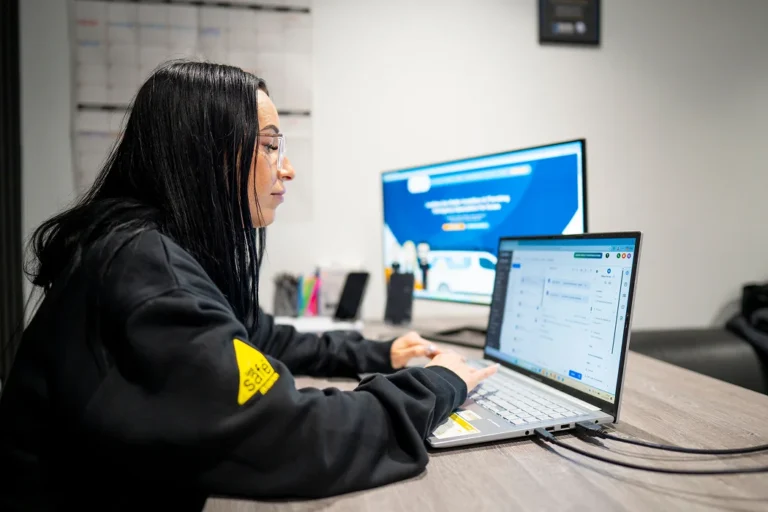 Woman working on laptop in an office setting, focused on digital tasks related to heating and plumbing services, with a calendar in the background.